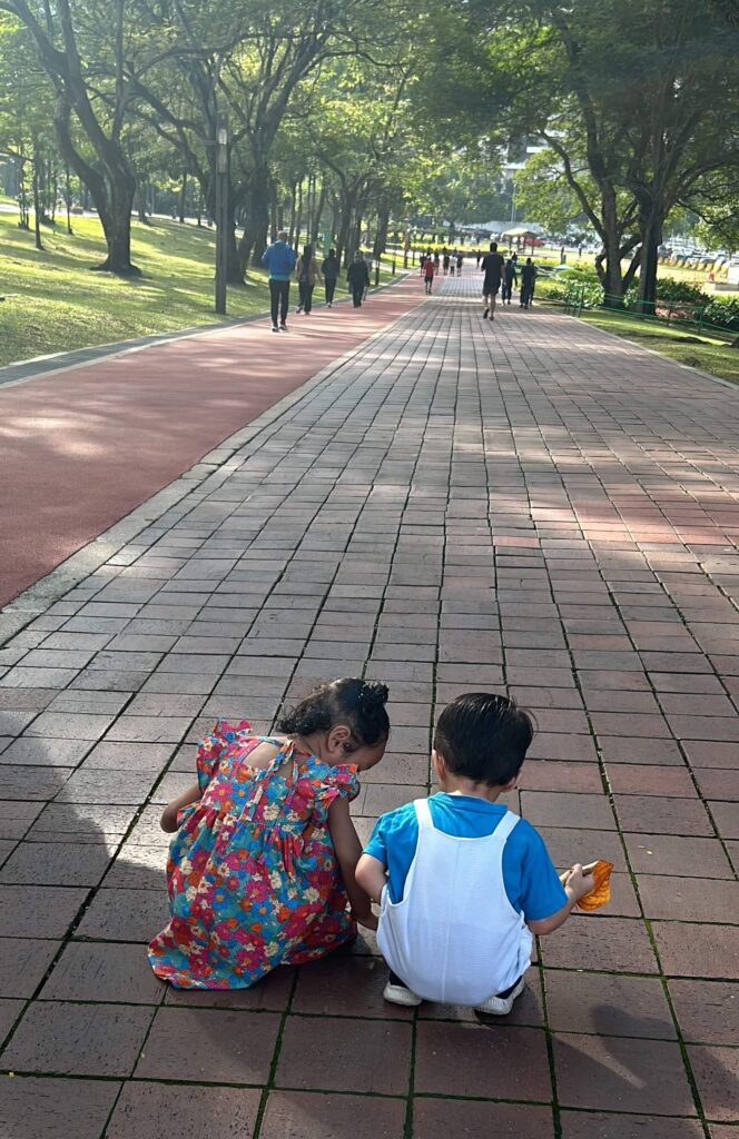 two kids playing together and crouching down on the ground along the jogging pathway at KLCC park 