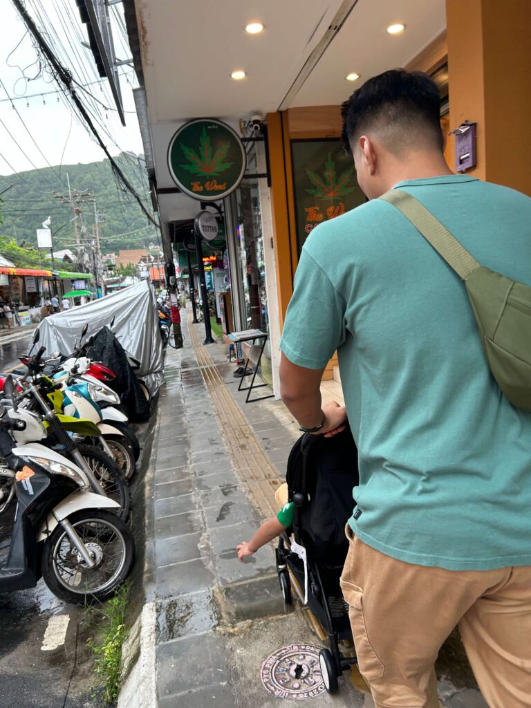 A dad pushing his son on a black stroller along the streets of Kata in Phuket 